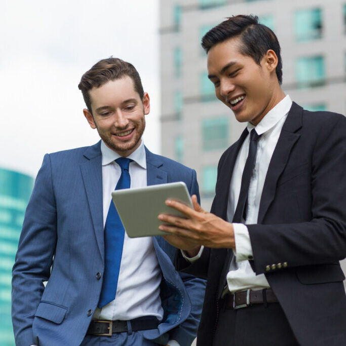 Closeup of Smiling Coworkers Using Tablet Outdoors Closeup of two smiling young business men using tablet computer and standing with office buildings in background